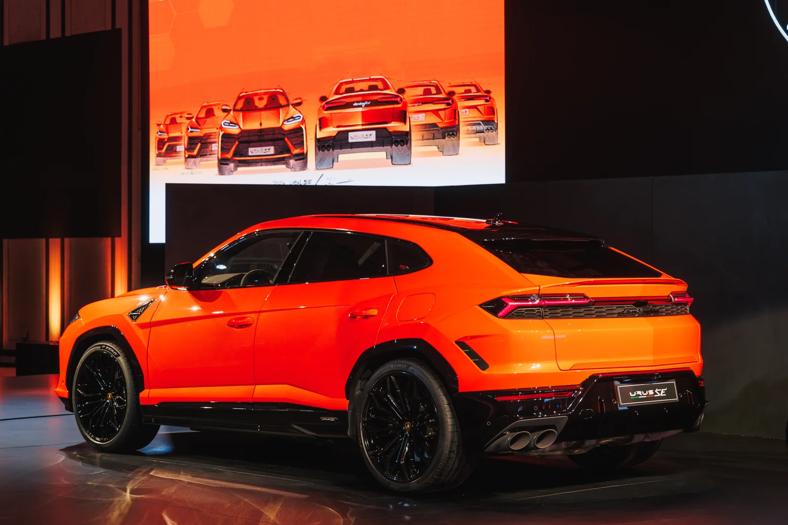 Rear three-quarter view of the orange Urus SE with design sketches on the screen behind