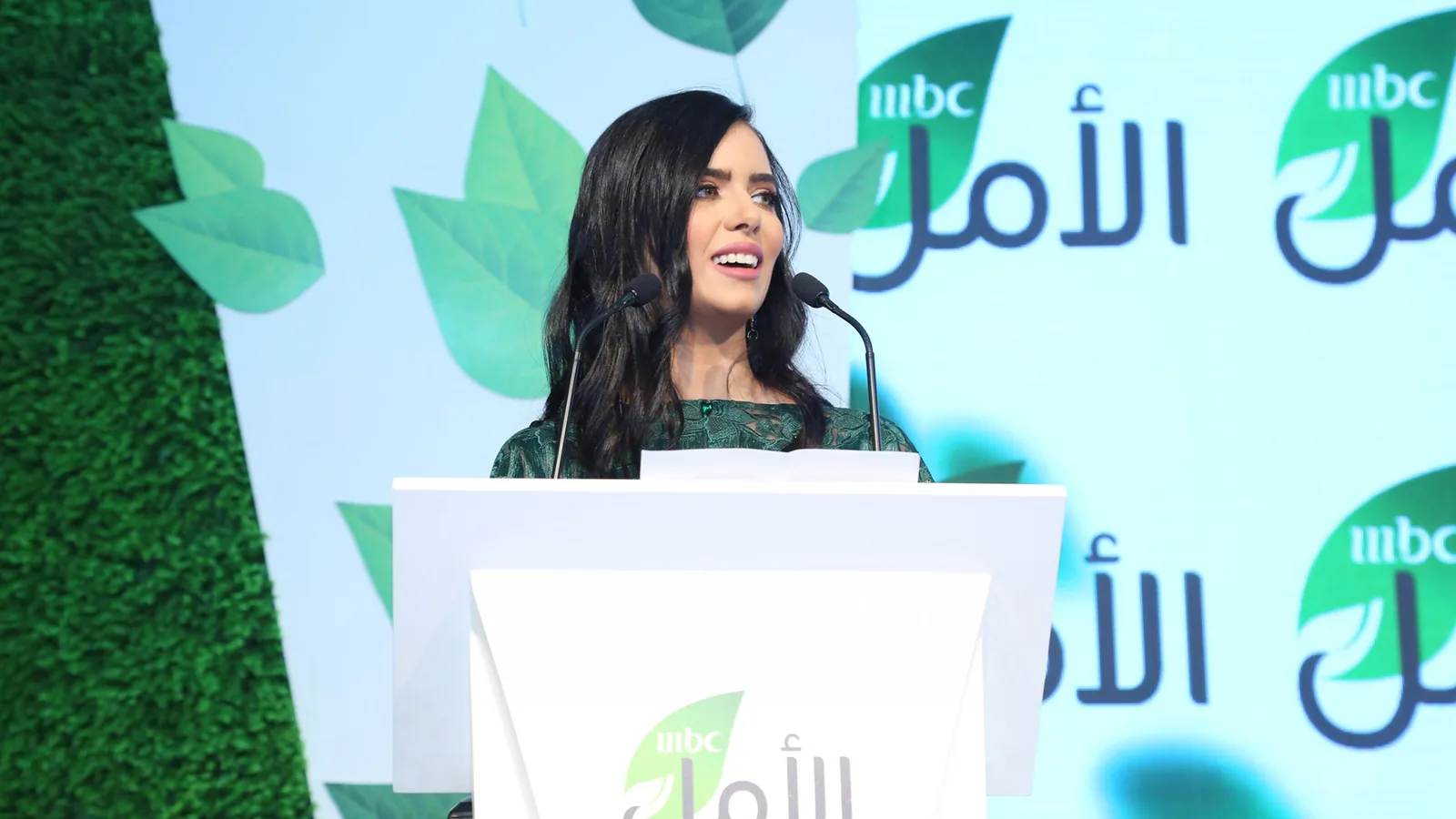 Host speaking at the podium in front of the MBC Al Amal logo